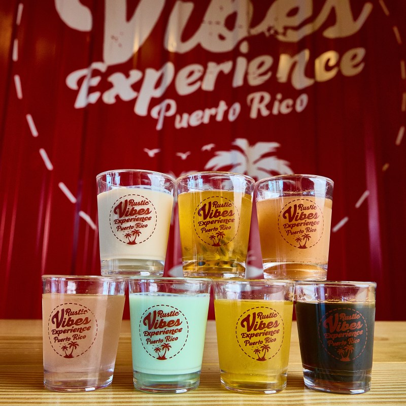 Seven colorful drink glasses with 'Rustic Vibes Experience Puerto Rico' logo on a wooden table.