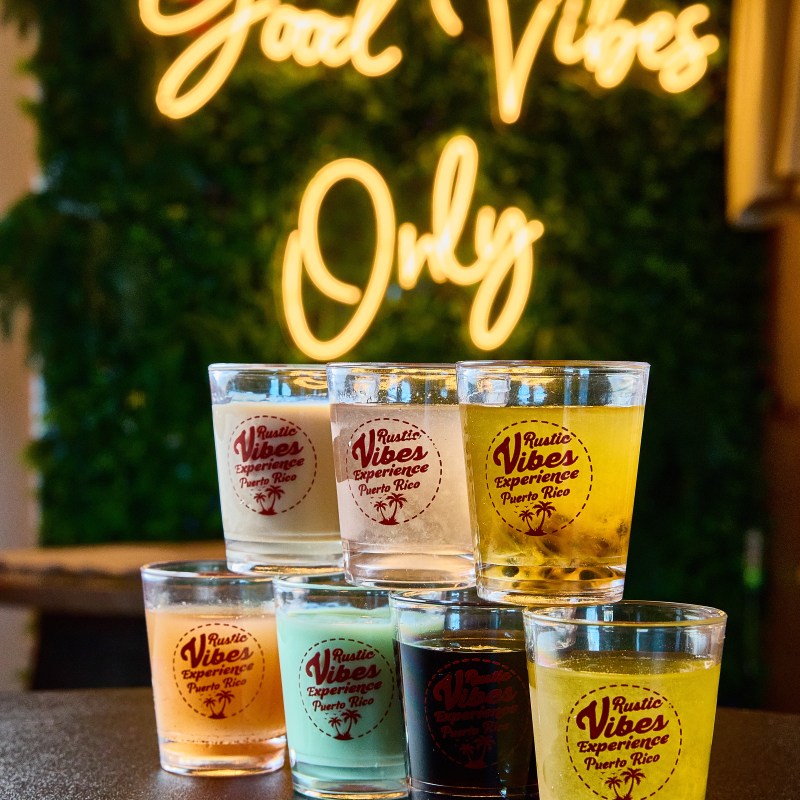Stacked colorful drinks in glasses, 'Good Vibes Only' neon sign in background.