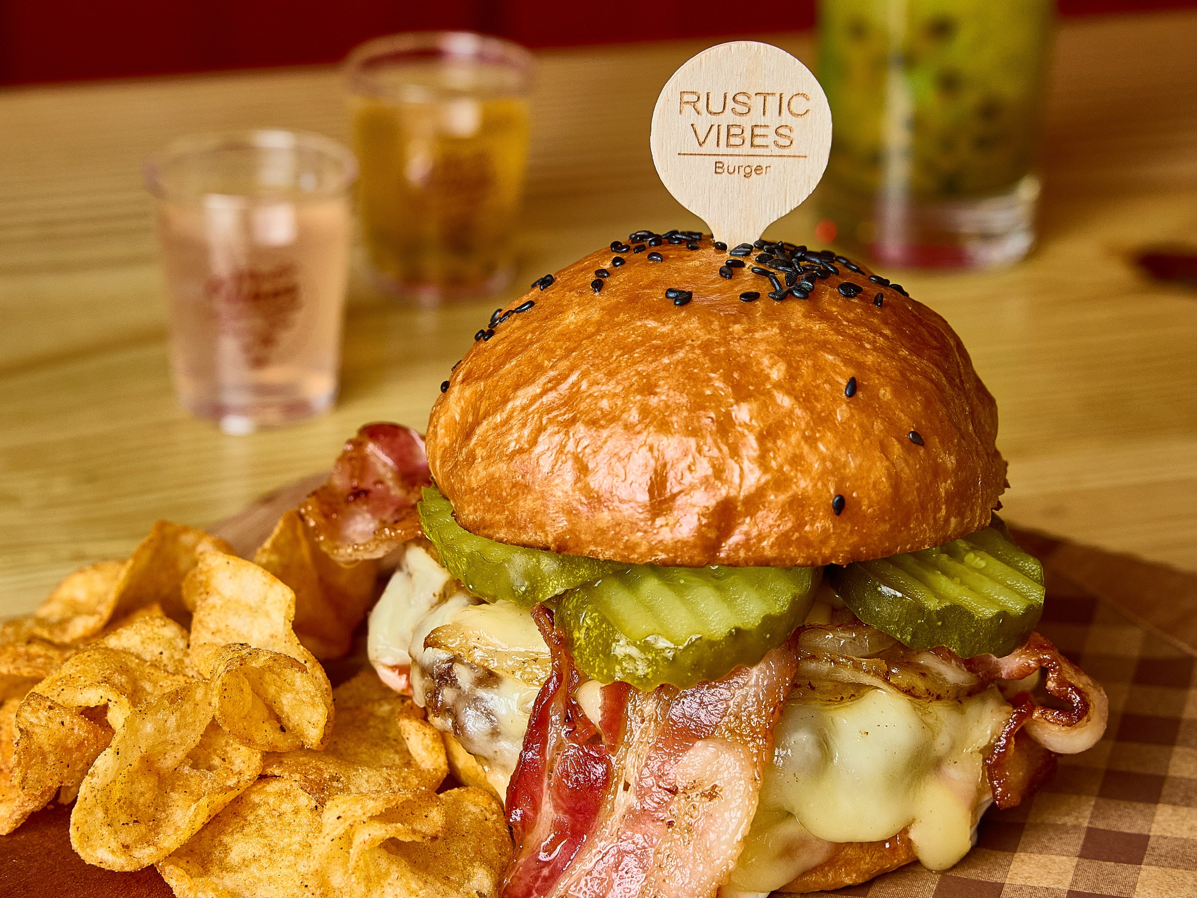 Cheeseburger with bacon, pickles, and chips on a wooden board with drinks in the background.