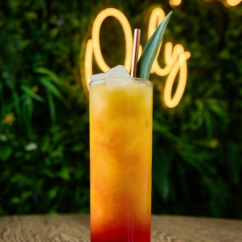 Tall colorful cocktail with ice and a leaf garnish on a wood table, neon sign in background.