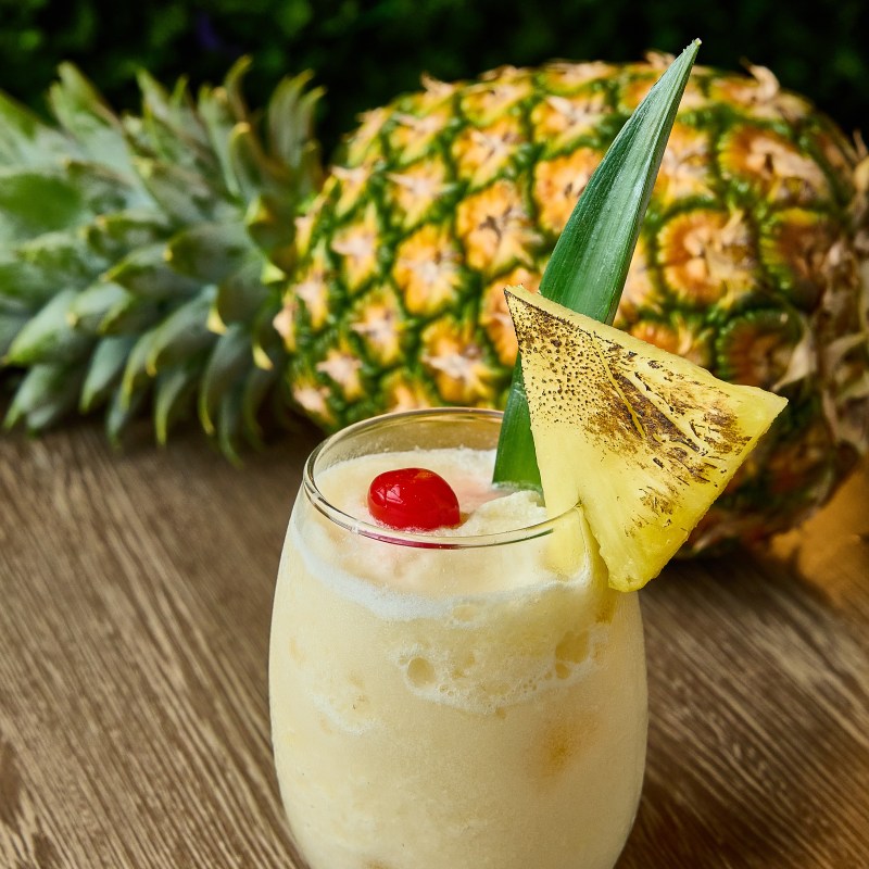 Pineapple cocktail in a glass with a cherry, pineapple slice, and leaf garnish, with a whole pineapple in the background.