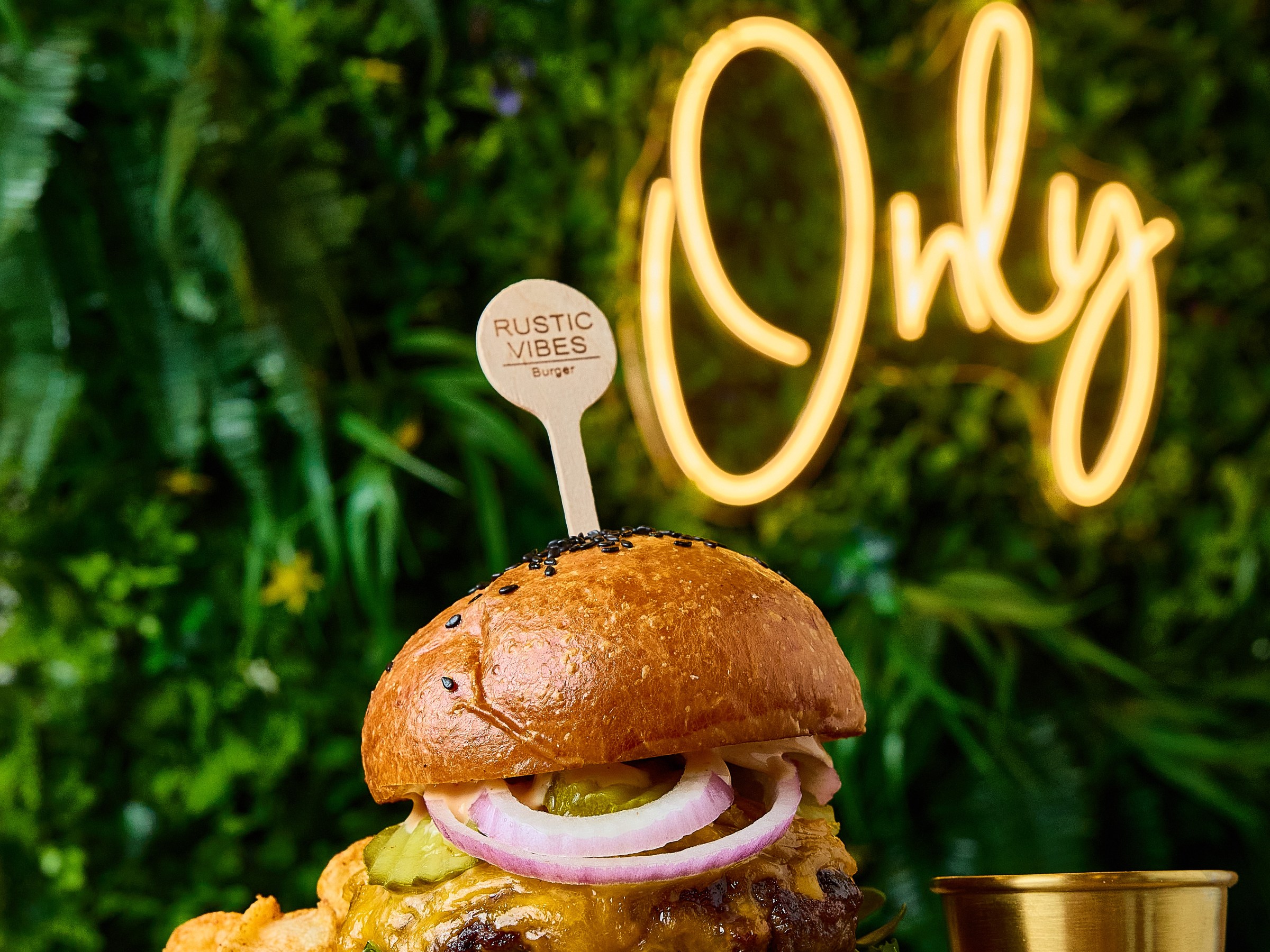 Burger with pickles and onions on a wooden board, neon 'Good Vibes Only' sign in the background.