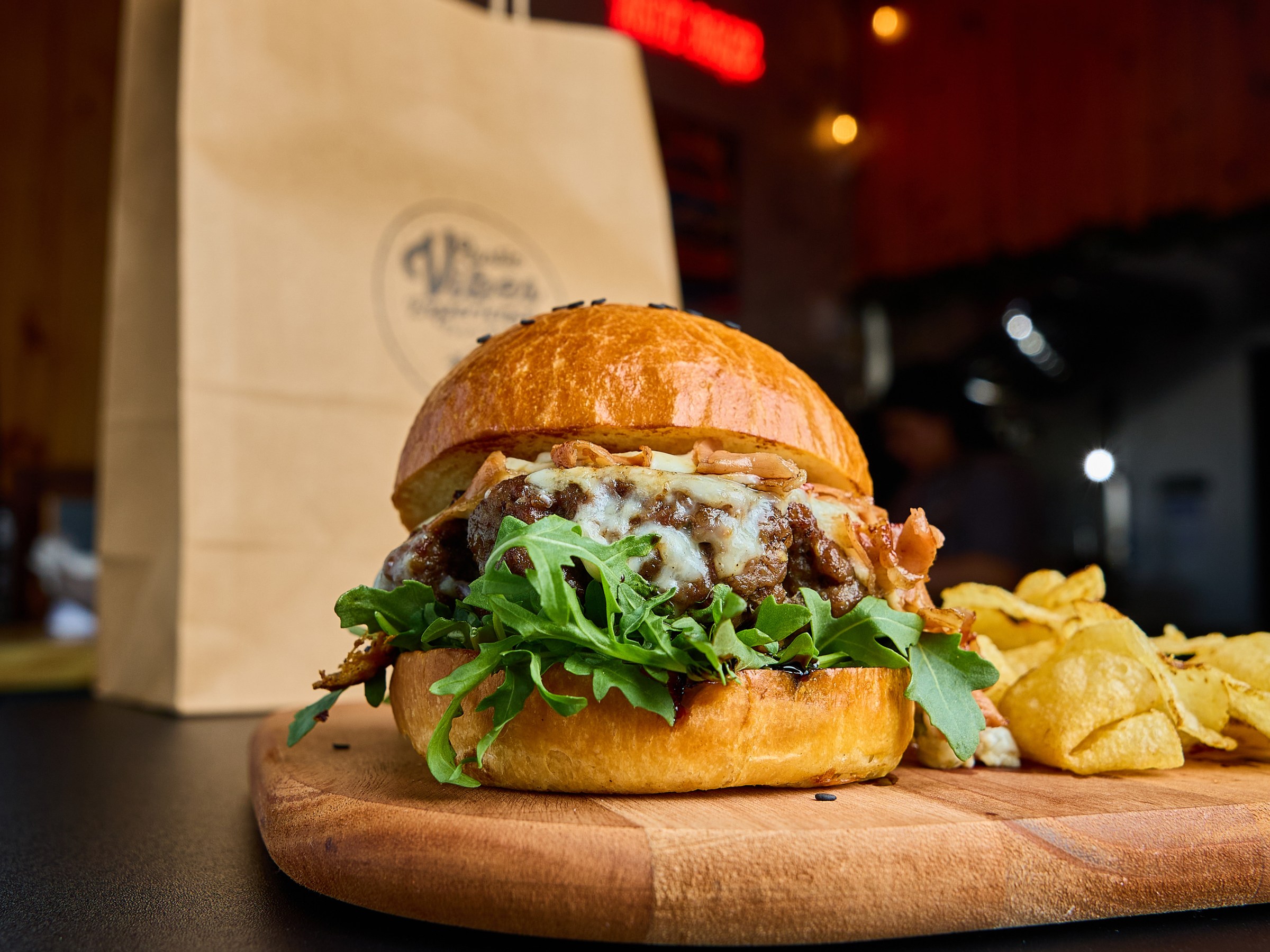 Gourmet burger with arugula and cheese on wooden board, chips side, paper bag background, warm lighting.