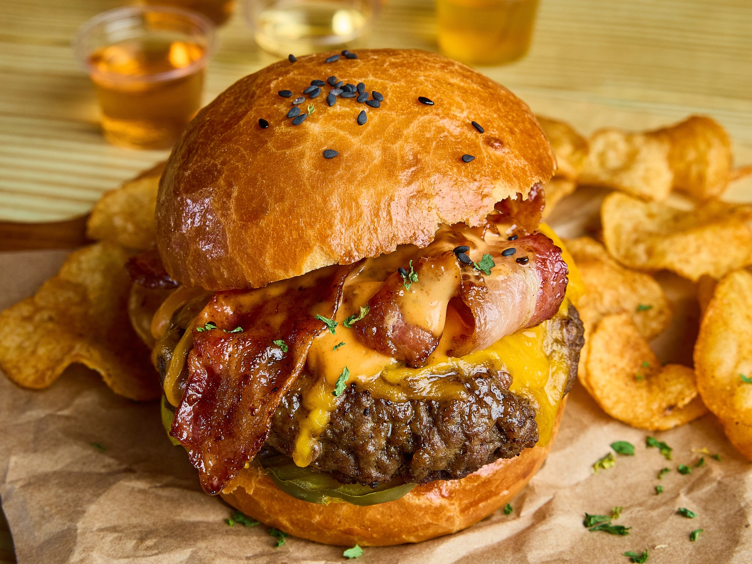 Burger with bacon, cheese on a bun, potato chips, drinks in background on a wooden table.