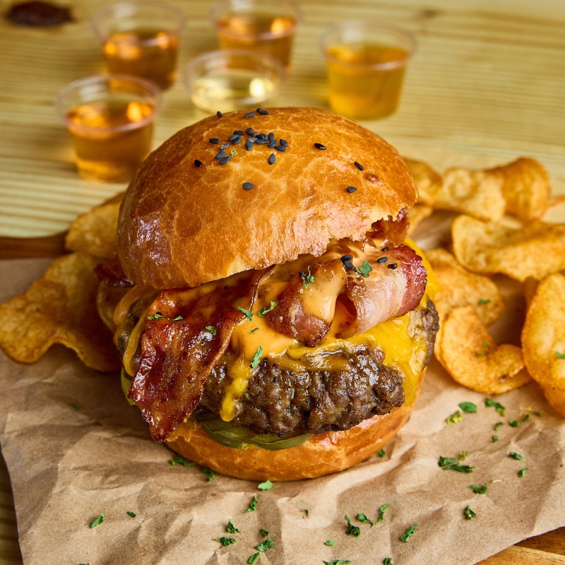 Burger with bacon and cheese on a bun, served with chips and drinks on the side.